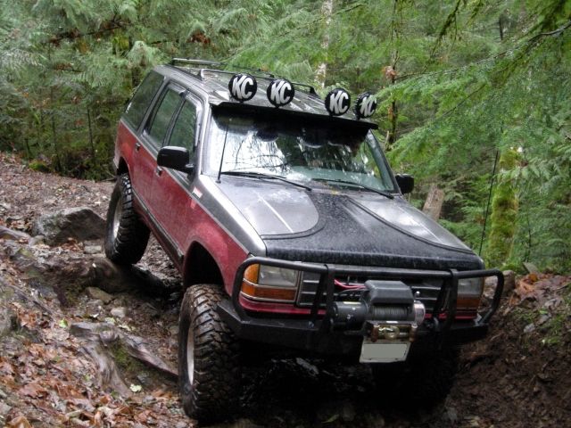 Red and black Ford Explorer off-roading on a rocky trail with winch and KC lights.