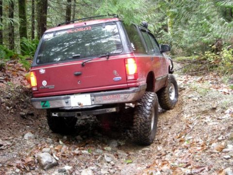 Red lifted Ford Explorer on a muddy forest trail, rear quarter view.