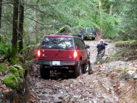 Red SUV on a muddy trail in a forest, with another SUV and a person in the background.