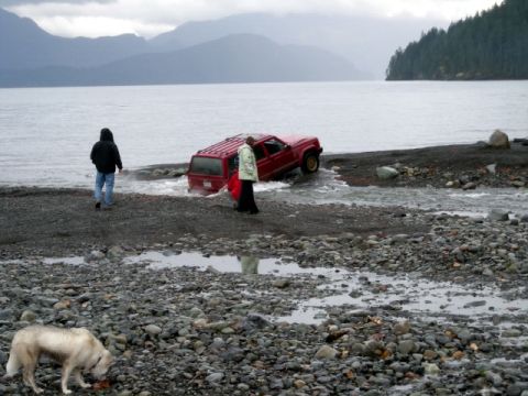 Red SUV stuck in water on a rocky beach with two people and a dog.