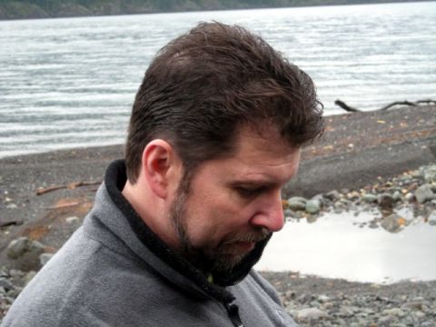 Man in gray fleece jacket looking down, with water and rocky shore in background.