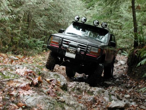 Black SUV with winch and KC lights on roof rack driving on a rocky, wooded trail.