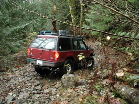 Red SUV driving on a rocky, muddy forest trail.