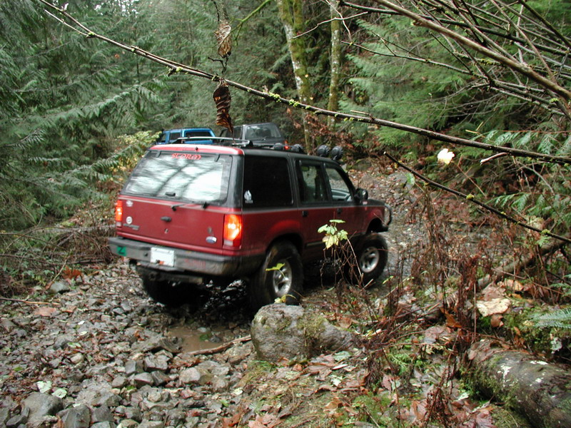 Red SUV driving on a rocky, muddy forest trail.