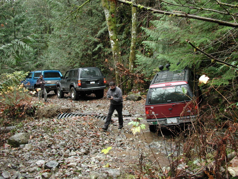 Three SUVs on a muddy, wooded trail, one red SUV stuck in water.