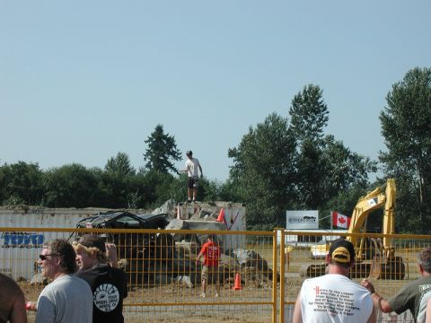 Person standing on rubble with wrecked vehicle and excavator in background.