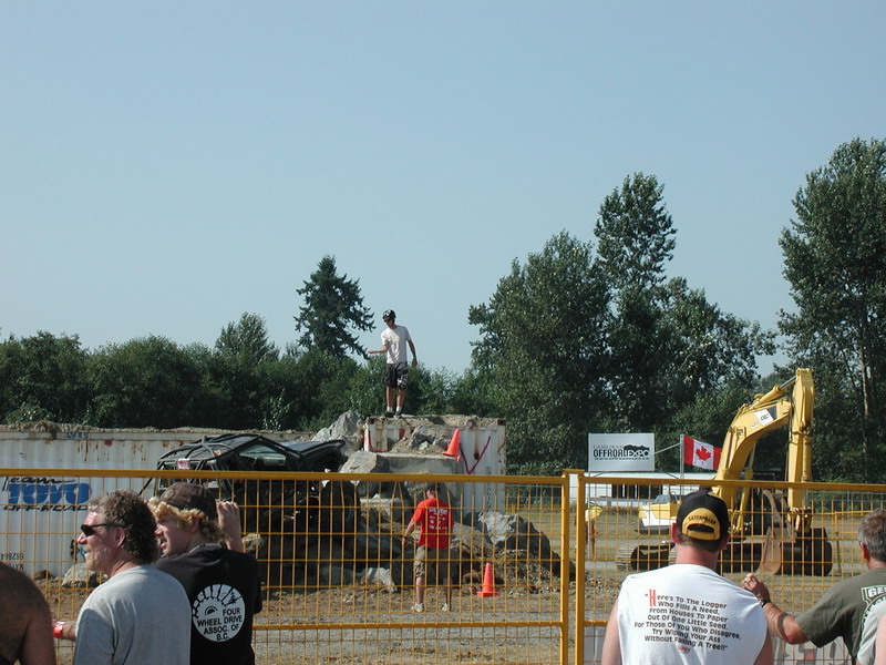 Person standing on rubble with wrecked vehicle and excavator in background.
