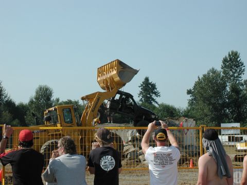 Black SUV being lifted by a yellow front loader, with spectators watching.