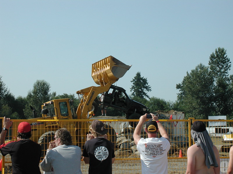 Black SUV being lifted by a yellow front loader, with spectators watching.