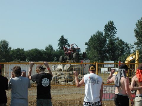 Red off-road buggy on a rocky obstacle course with spectators behind a fence.