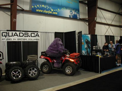 Purple mascot on a red ATV with stacks of tires and a QUADS.CA banner.