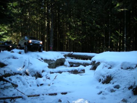 1985 Ford Bronco - Two off-road vehicles navigate a snow-covered forest trail with fallen logs and rocky terrain. The lead vehicle, a light-colored SUV, is po…