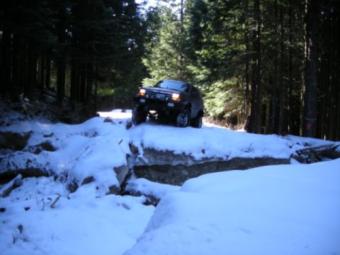 1985 Ford Bronco - A dark-colored SUV drives through a snowy, wooded area, navigating over rocks and snow drifts on a trail.