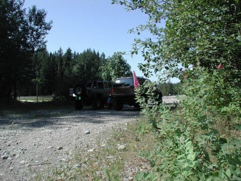 1985 Ford Bronco - Two off-road vehicles are parked on a gravel road surrounded by trees. The vehicle in the foreground is a dark-colored SUV with its spare t…