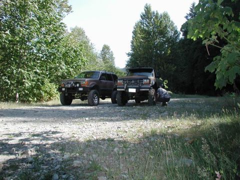 1985 Ford Bronco - Two lifted off-road vehicles, a dark brown SUV and a dark brown pickup truck, are parked on a gravel road surrounded by trees. A person is…