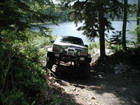 1985 Ford Bronco - A modified 1985 Ford Bronco is navigating a rocky, wooded trail next to a lake. The vehicle features large tires, a winch, and auxiliary li…