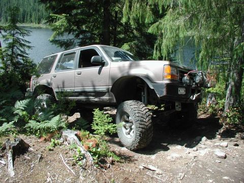 1985 Ford Bronco - A lifted, tan Ford Explorer SUV is parked off-road next to a lake, surrounded by trees and ferns. The vehicle features large, aggressive ti…