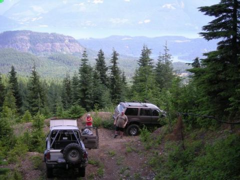 1985 Ford Bronco - Two off-road vehicles are parked on a dirt path with a scenic mountain and lake view in the background. A modified pickup truck with a roll…
