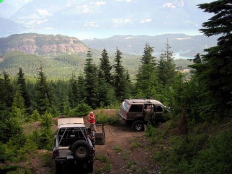 1985 Ford Bronco - Two off-road vehicles are parked on a dirt trail overlooking a vast, forested valley with a body of water in the distance. One vehicle is a…