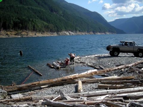1985 Ford Bronco - A lifted, light-colored pickup truck is parked on a rocky shore next to a large body of water, with two people relaxing near the water's ed…