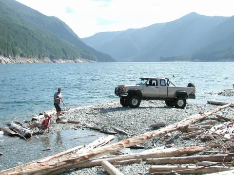 1985 Ford Bronco - A lifted, light-colored pickup truck is parked on a rocky shore next to a large body of water with mountains in the background. Two people…