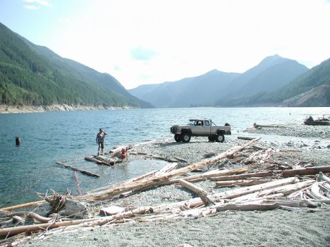 1985 Ford Bronco - A light-colored pickup truck is parked on a rocky shore next to a large body of water. A person stands in the water near the shore, and ano…