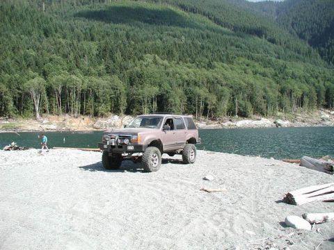 1985 Ford Bronco - A lifted, modified SUV is parked on a gravelly shore next to a body of water with a forested mountain in the background.