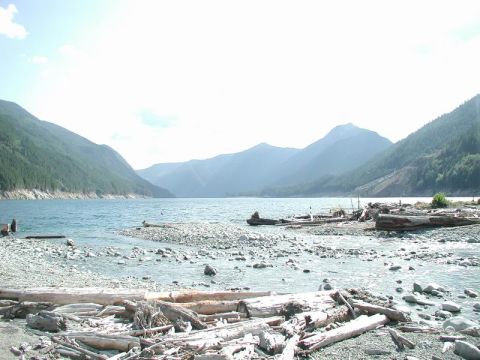 1985 Ford Bronco - A wide shot of a lake surrounded by mountains, with a rocky shore in the foreground littered with driftwood.