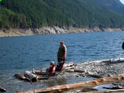 1985 Ford Bronco - Two people sit in camping chairs on a rocky shore next to a body of water, with a forested mountainside in the background. A man stands beh…
