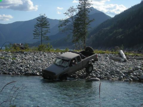 1985 Ford Bronco - A gray pickup truck is partially submerged in a shallow, rocky river, with its front tires on the riverbed and its rear tires on the bank.…