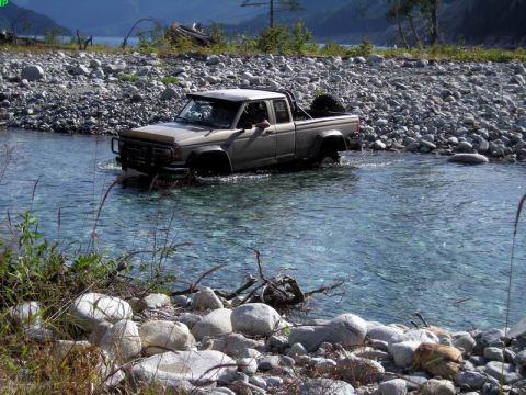 1985 Ford Bronco - A tan pickup truck drives through a shallow, rocky river. The truck has a spare tire mounted on the back and appears to be an older model.