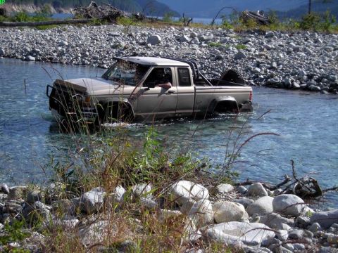 1985 Ford Bronco - A tan 4-door pickup truck is driving through a shallow, clear river with a rocky shore and sparse vegetation in the foreground. The truck h…