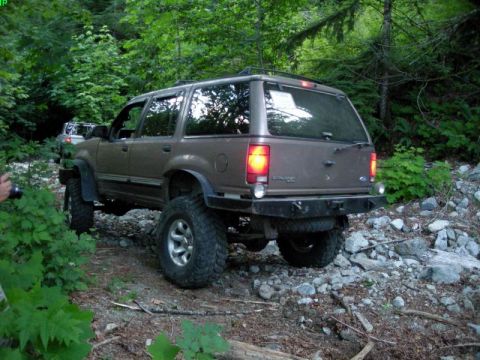 1985 Ford Bronco - A brown Ford Explorer SUV with off-road tires is driving on a rocky trail through a forest. Another vehicle is visible in the background.