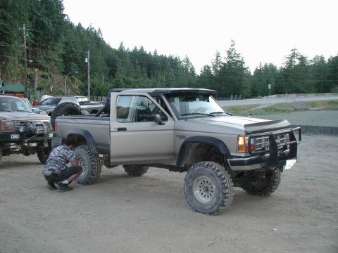 1985 Ford Bronco - A lifted, tan Ford Ranger pickup truck with large off-road tires is parked on a dirt lot. A person in camouflage clothing is kneeling by th…