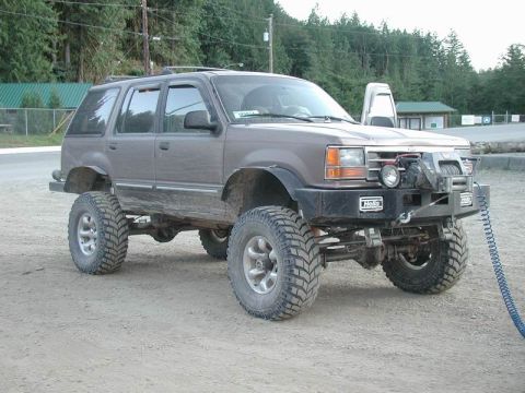 1985 Ford Bronco - A lifted brown 1990s Ford Explorer SUV with large off-road tires and a winch mounted on the front bumper is parked on a dirt lot.