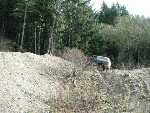 1985 Ford Bronco - A dark-colored SUV is driving up a steep, rocky incline in a wooded area. The vehicle appears to be a Ford Bronco, possibly from the mid-19…