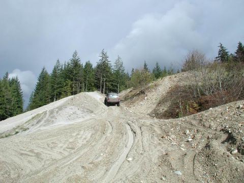 1985 Ford Bronco - A dark colored SUV drives up a steep, loose dirt and gravel hill. Tall evergreen trees line the top of the hill under a cloudy sky.