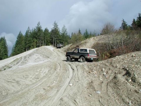 1985 Ford Bronco - A dark-colored Ford Bronco with a white camper shell drives up a steep, sandy hill on a dirt road. Another SUV is visible further up the in…