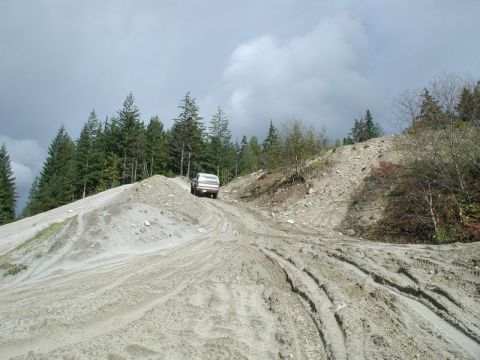 1985 Ford Bronco - A white and red Ford pickup truck drives up a steep, muddy, and sandy incline on a dirt road. The truck is surrounded by trees and a cloudy…