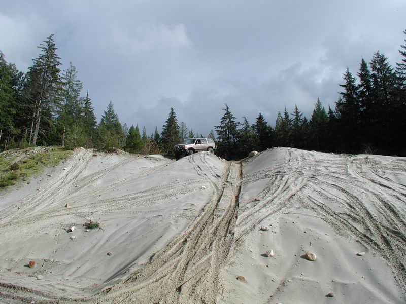 Light-colored SUV driving up a sandy hill with tire tracks, evergreen trees in background.