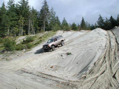 1985 Ford Bronco - A silver SUV is driving down a steep, sandy hill with tire tracks visible on the surface. Evergreen trees line the background of the scene.