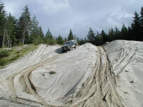 1985 Ford Bronco - A dark-colored SUV is driving up a steep, sandy incline with numerous tire tracks visible. The vehicle is positioned on the right side of t…