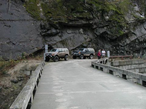 1985 Ford Bronco - Two modified SUVs are parked on a concrete bridge next to a rocky cliff face. A brown SUV is on the left and a black Bronco is to its right…