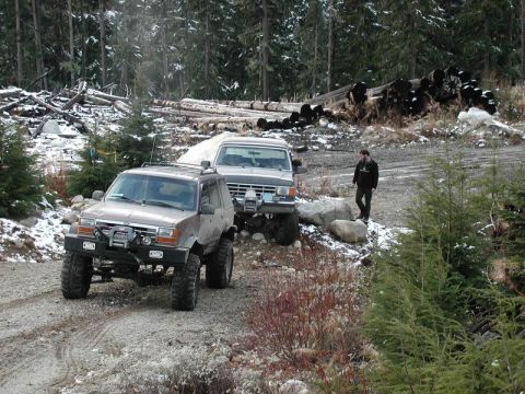 1985 Ford Bronco - Two off-road modified SUVs are parked on a gravel road in a wooded area with snow on the ground. A person stands near the second SUV, which…
