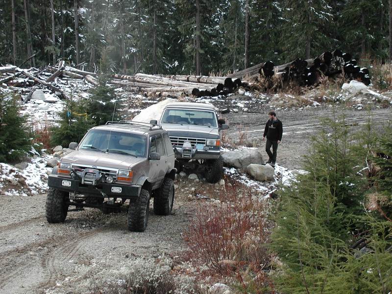 Two modified SUVs on a gravel road in a snowy, wooded area. A person stands near the second SUV.
