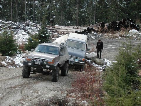 1985 Ford Bronco - Two modified SUVs are navigating a rough, wooded terrain with snow on the ground. A person stands on a rock next to the second SUV, observi…