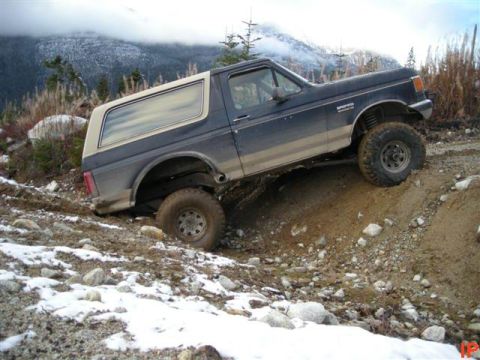 1985 Ford Bronco - A dark blue Ford Bronco is navigating a steep, muddy, and rocky incline with patches of snow. The vehicle is angled upwards, with its front…
