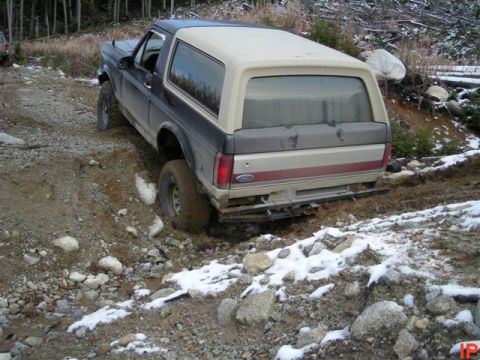 1985 Ford Bronco - A dark blue and tan Ford Bronco is navigating a muddy, rocky trail with snow patches. The rear passenger side wheel is deep in a rut, indic…