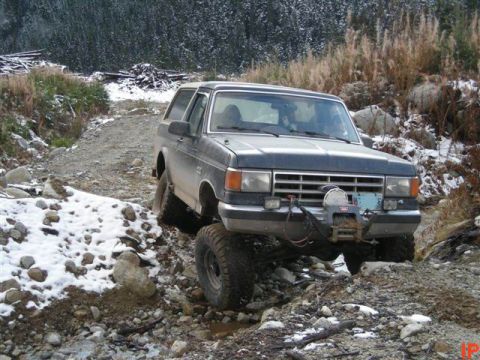 1985 Ford Bronco - A dark colored Ford Bronco is driving on a rocky, snowy, off-road trail. The vehicle has a winch mounted on the front bumper.