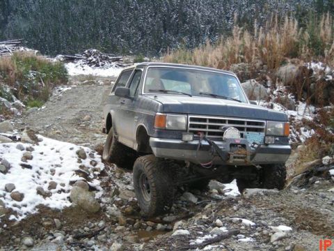 1985 Ford Bronco - A dark-colored Ford Bronco is navigating a rocky, off-road trail with patches of snow. The vehicle is equipped with a winch mounted on the…